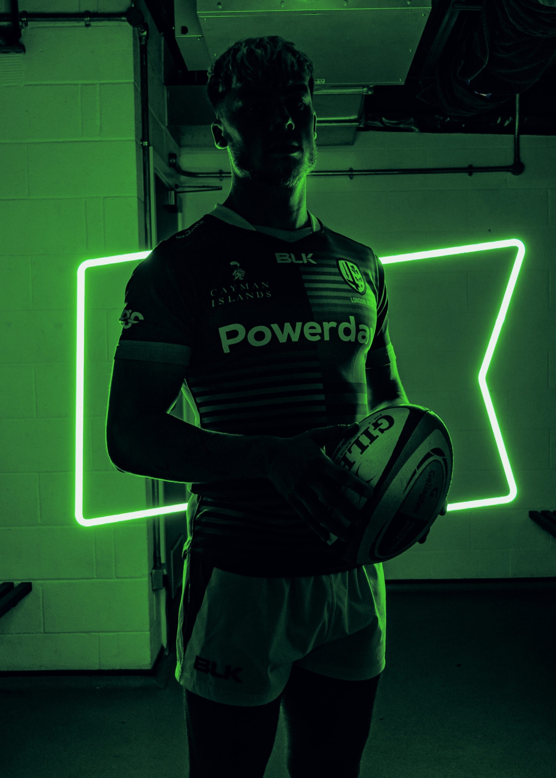 A deeply shadowed and square-jawed man in team kit and holding a rugby ball backlit by a neon green outline of a flag